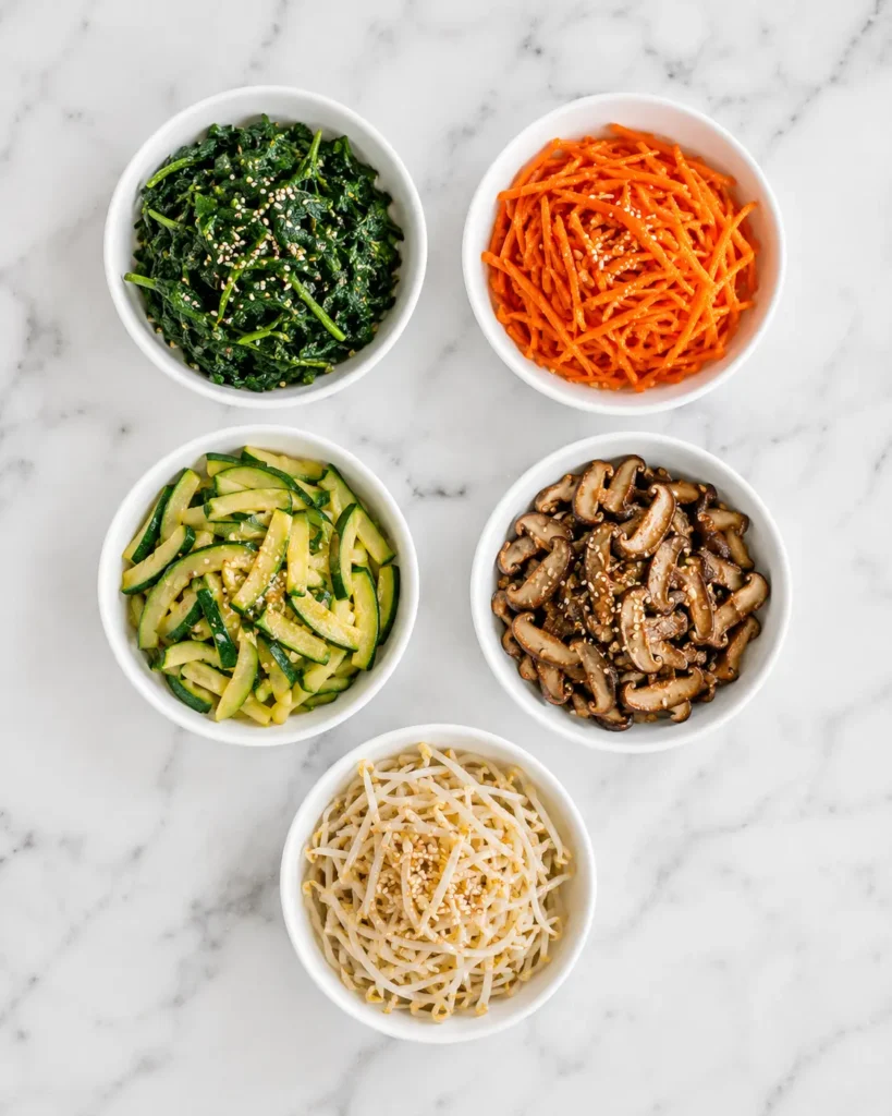 Five bowls of seasoned vegetables for bibimbap including spinach, carrots, zucchini, mushrooms, and bean sprouts